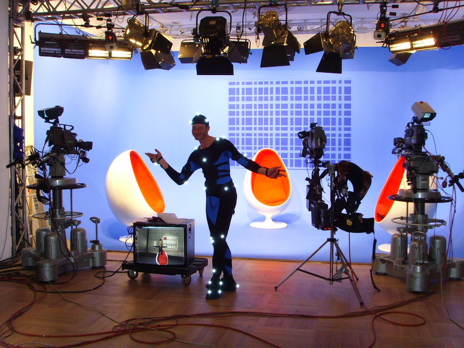 A performer in a marker suit in the Theater im Zimmer bluebox, with Hugo the troll shown live in a virtual set on the output monitor beside him