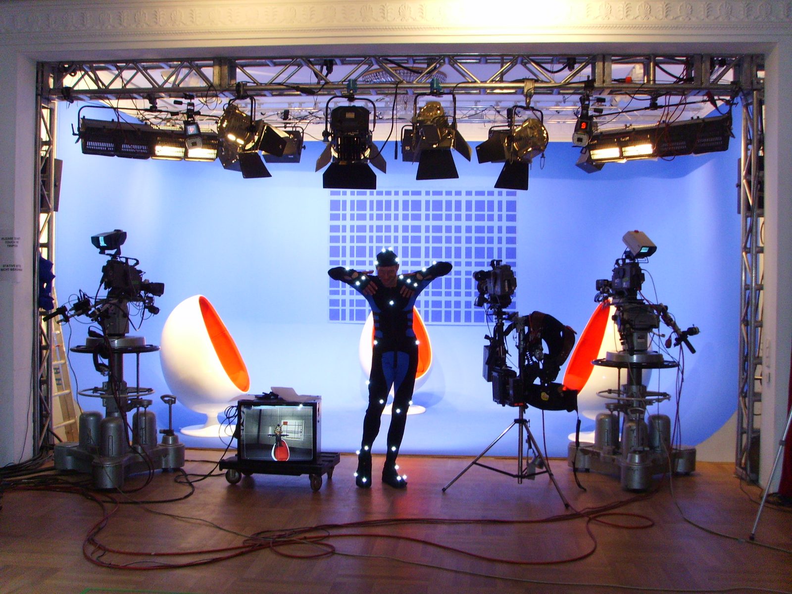 Wide view of the virtual studio on the theatre stage: performer in marker suit, tracked cameras, output monitor, beneath the theatre's stucco cornice